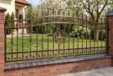Kute ogrodzenie posesji w stylu klasycznym z ornamentyką florystyczną, model OK57 — aranżacja: kremowy dom z ceglaną podmurówką i sadem ogrodowym. Wizualizacja AI.