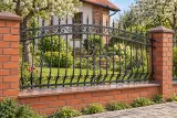 Kute ogrodzenie posesyjne w stylu klasycznym z ornamentyką spiralną, model mg21 — aranżacja: dom z cegły i tynku z sadem i kamiennym chodnikiem. Wizualizacja AI.