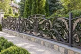 Kute ogrodzenie posesyjne z ornamentyką florystyczną w estetyce barokowej, model mg13 — aranżacja: zaciszna posesja parkowa z dojrzałymi iglakami i żwirową ścieżką. Wizualizacja AI.