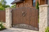 Kute ogrodzenie posesyjne z panelem drewnianym i ornamentyką florystyczną, model mg19 — aranżacja: drewniany dom z jasnymi słupkami wapiennymi i ścieżką ogrodową. Wizualizacja AI.