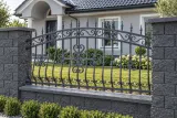 Kute ogrodzenie rezydencjalne z zdobionymi przęsłami stylu klasycznego, model mg24 — aranżacja: jasnoszara willa z ciemnymi słupkami kamiennymi i ogrodem. Wizualizacja AI.