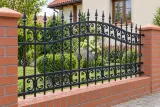 Kute przęsło ogrodzeniowe tradycyjne z ornamentyką spiralną, model OK11 — aranżacja: dom z cegły i tynku z uporządkowaną rabatą bylinową. Wizualizacja AI.