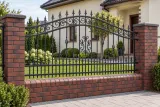 Kute przęsło ogrodzeniowe z dekoracyjnymi spiralami i grotami, model OK33 — aranżacja: spokojna posesja z kremowym domem i ciemnymi słupkami ceglanymi. Wizualizacja AI.