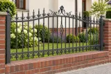 Kute przęsło ogrodzeniowe z ornamentalnym zdobieniem, model OK05 — aranżacja: klasyczny dom z ceglaną podmurówką i rabatą bylinową. Wizualizacja AI.