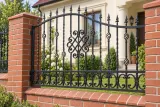 Kute przęsło ogrodzeniowe z ornamentyką klasyczną i akcentami spiralnymi, model OK60 — aranżacja: klasyczny kremowy dom z ceglanymi słupkami i ogrodem. Wizualizacja AI.