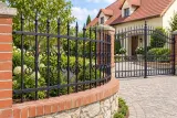 Kute przęsło ogrodzeniowe z ornamentyką klasyczną i fiolkami, model OK10 — aranżacja: łagodny podjazd przy domu z ceglano-kamienną podmurówką. Wizualizacja AI.