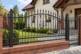 Ogrodzenie kute stalowe klasyczne z elementami ornamentalnymi, model OK51 — aranżacja: kremowy dom z drewnianymi detalami dachu i ceglaną podmurówką. Wizualizacja AI.
