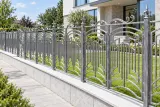 Ornamentalne kute ogrodzenie stalowe, model mg06 — aranżacja: współczesna rezydencja z kamienia i szkła z szarą podmurówką. Wizualizacja AI.